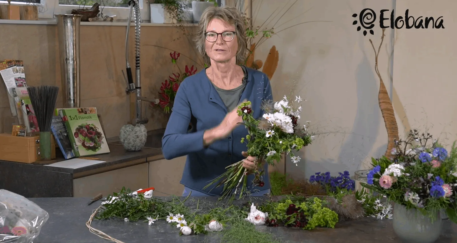Eleonore Schick erklärt Blumengesteck vor Floristik-Werkstatt mit Elobana-Logo