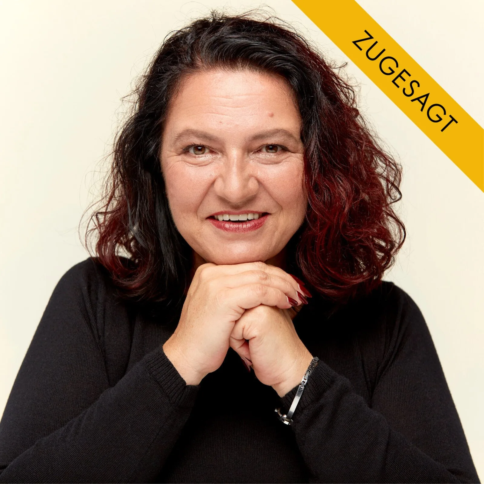 Brigitte Kraxner, Frau mit dunkelrotem lockigem Haar, schwarzem Pullover, Hände vor der Brust, vor hellem Hintergrund