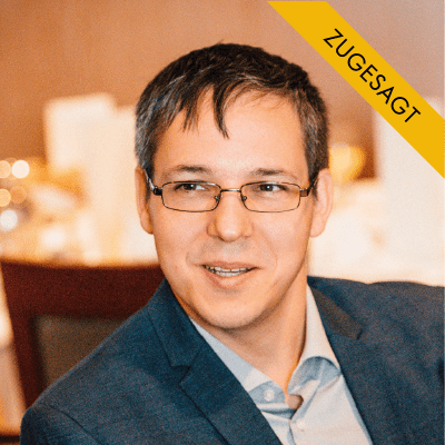 Daniel Förster, Mann mit dunklem Haar, Brille, dunklem Blazer und hellblauem Hemd, vor unscharfem Hintergrund
