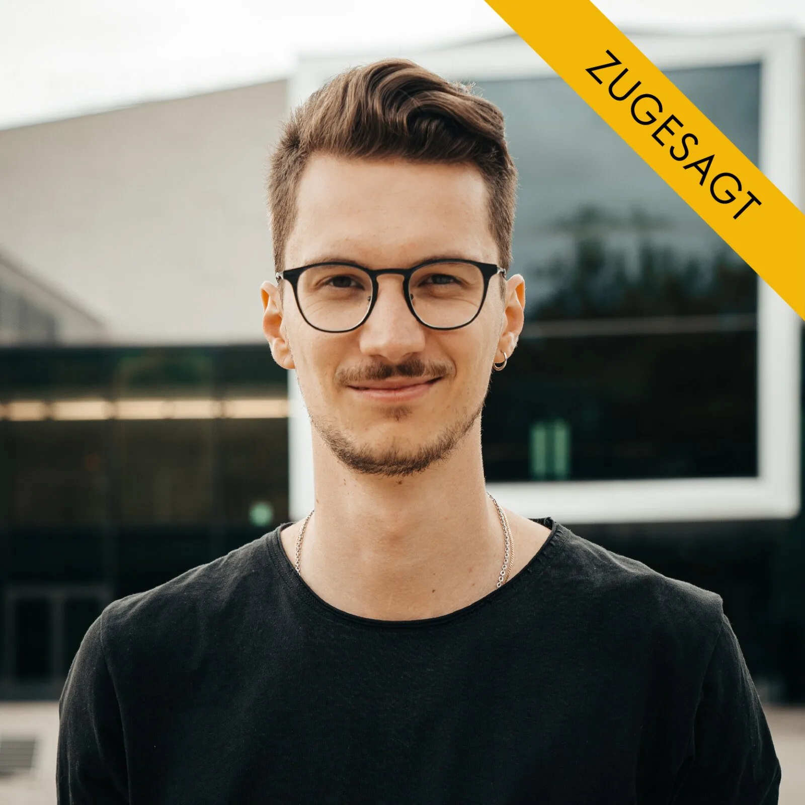 David Hollerer, junger Mann mit dunklem Haar, Brille, dunklem T-Shirt mit Kette, vor modernem Hintergrund