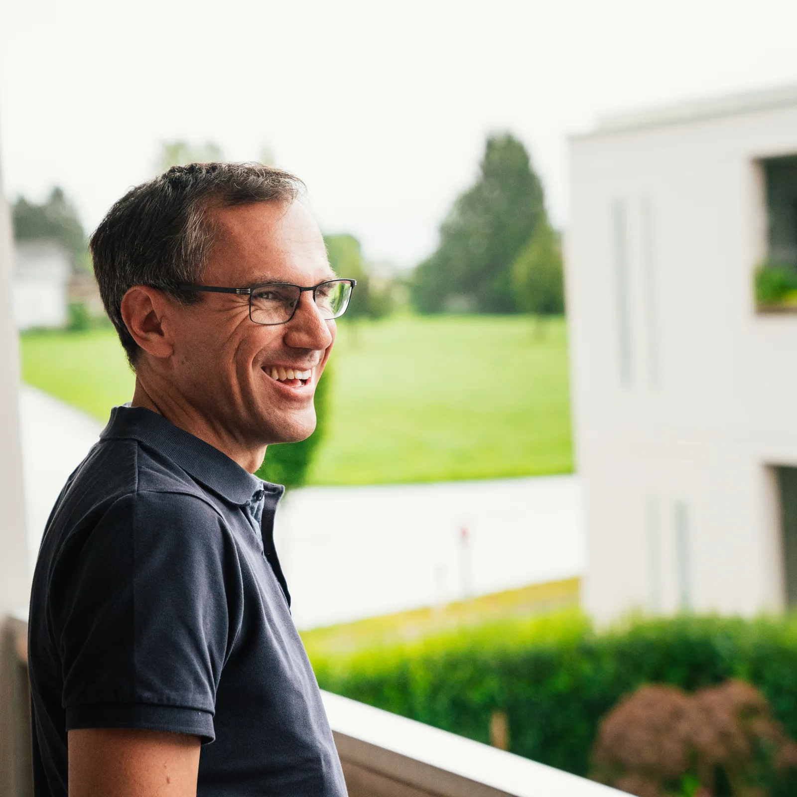 Thomas Fuchs lächelt in die Kamera, trägt Brille und dunkles Poloshirt, Garten und Haus im Hintergrund