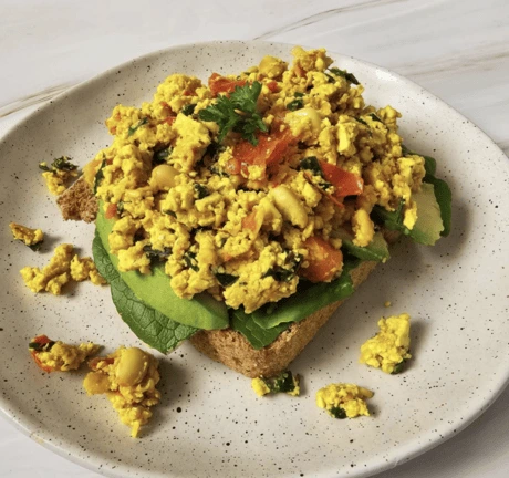 Tofu-Rührei auf Avocado-Toast mit Tomaten und Kräutern auf weißem Teller