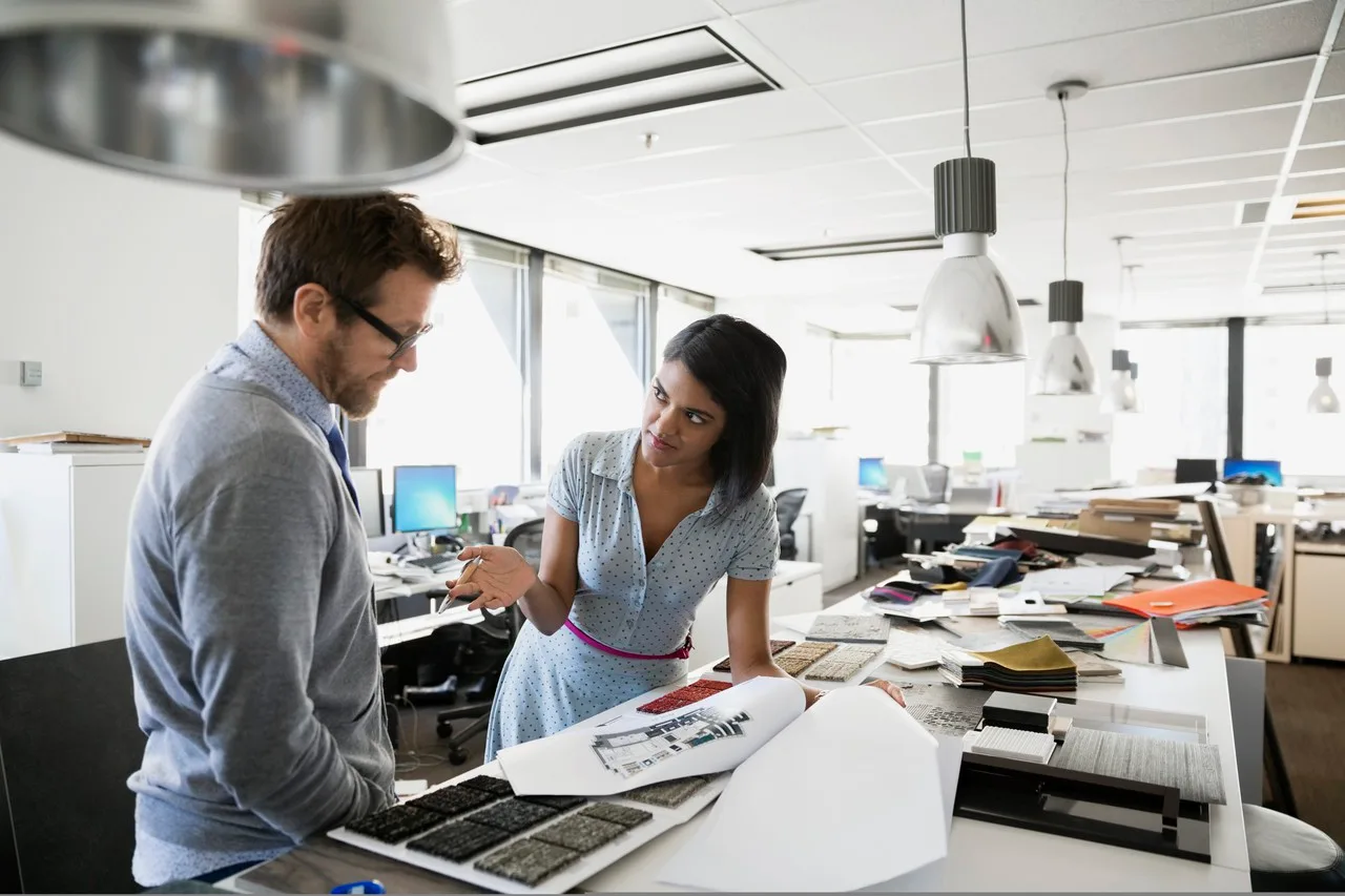 Mann und Frau besprechen Farbmuster und Materialien an Schreibtisch in hellem Büro