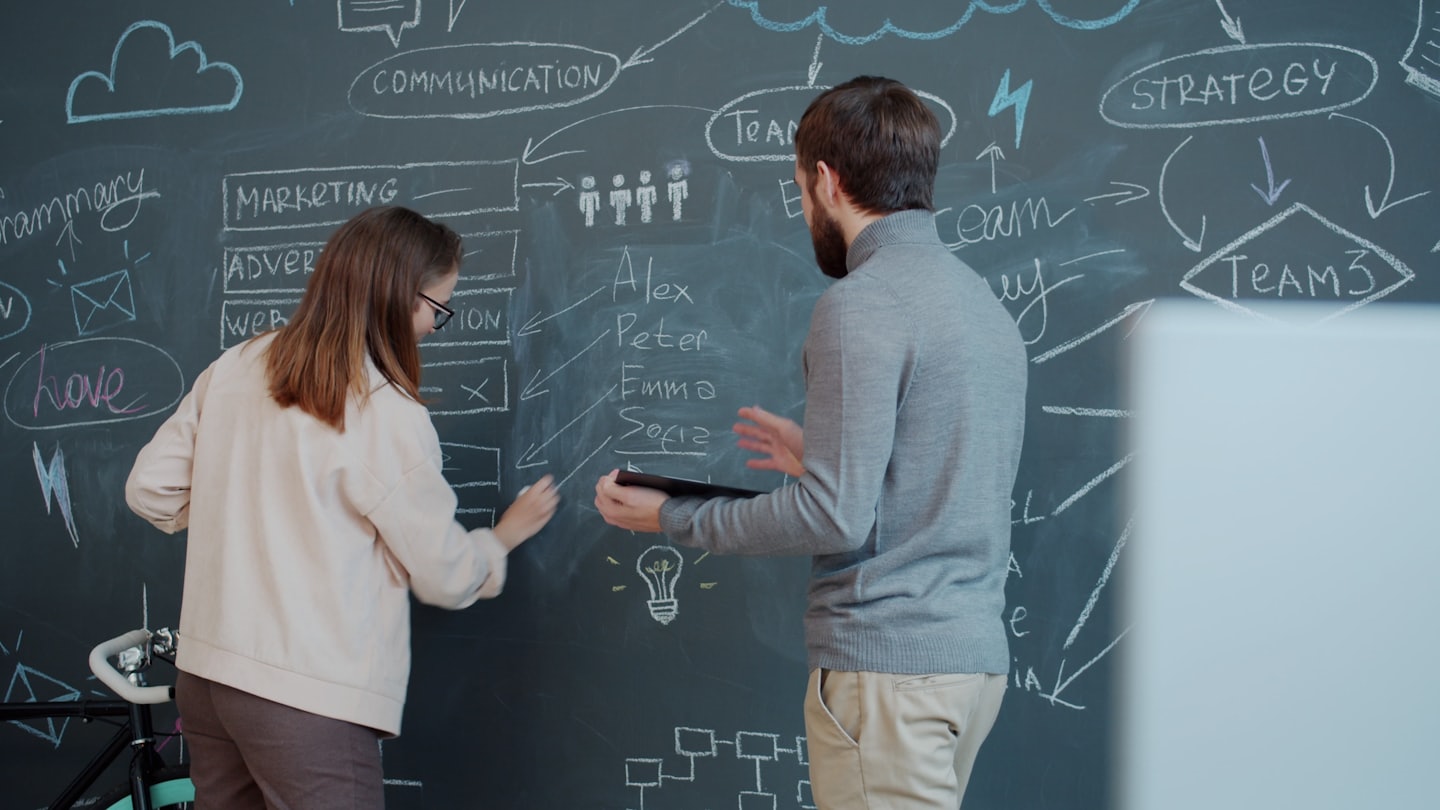 Zwei Personen vor Tafel mit Geschäftskonzept-Skizzen: Marketing, Advertising, Communication, Team, Strategy