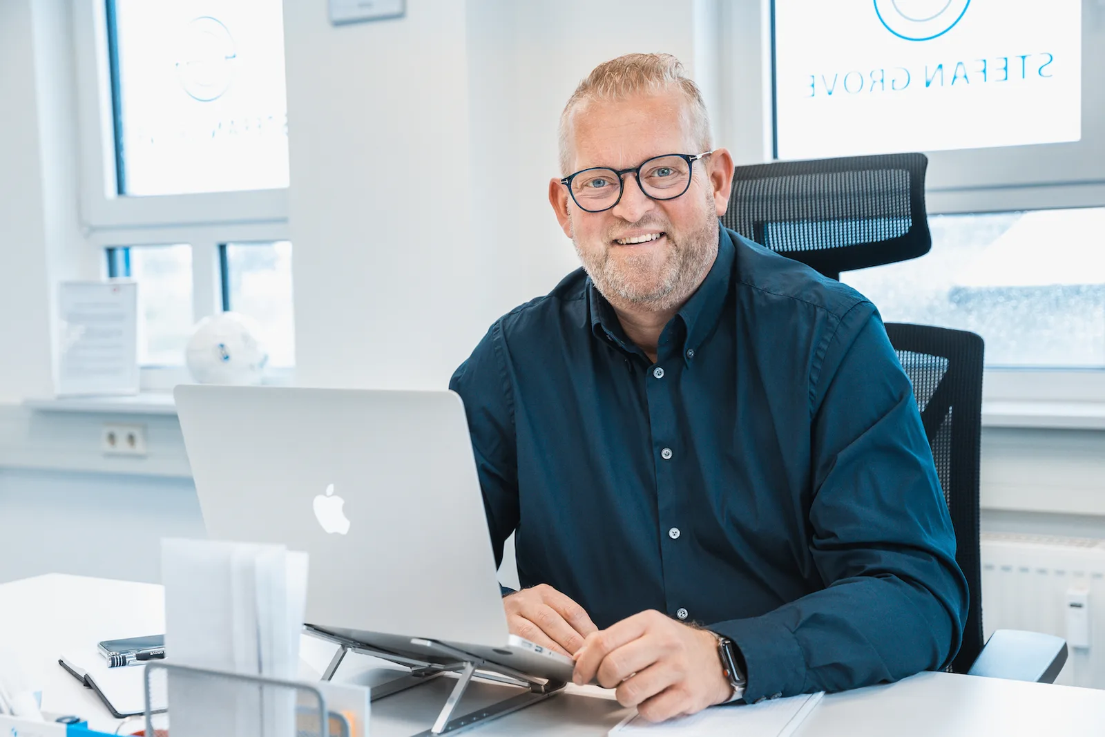 Stefan Grove sitzt am Schreibtisch im hellen Büro mit Laptop und lächelt in die Kamera