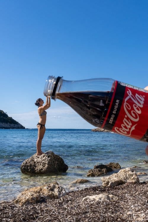 Person hält übergroße Coca-Cola-Flasche am Strand, Perspektivtrick