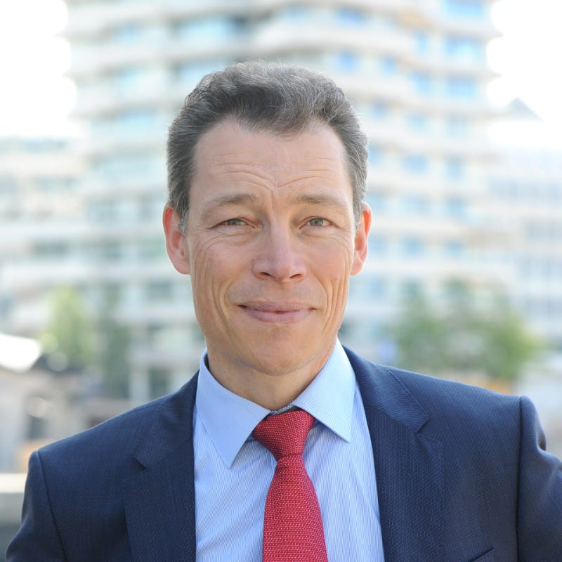 Porträt von Martin Wehrle in Anzug und roter Krawatte vor unscharfem Stadtgebäude-Hintergrund
