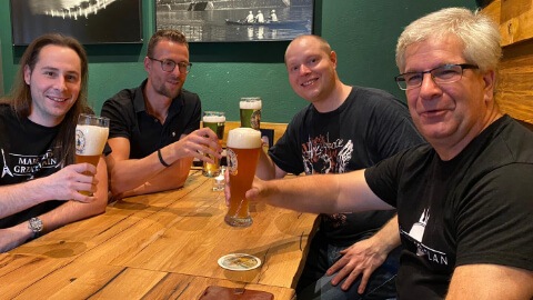Vier Männer sitzen an Holztisch mit Bier, entspannte Atmosphäre in grünem Raum