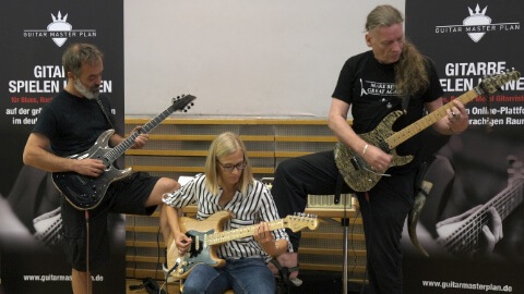 Drei Gitarristen spielen zusammen auf einer Bühne mit Guitar Master Plan Logo