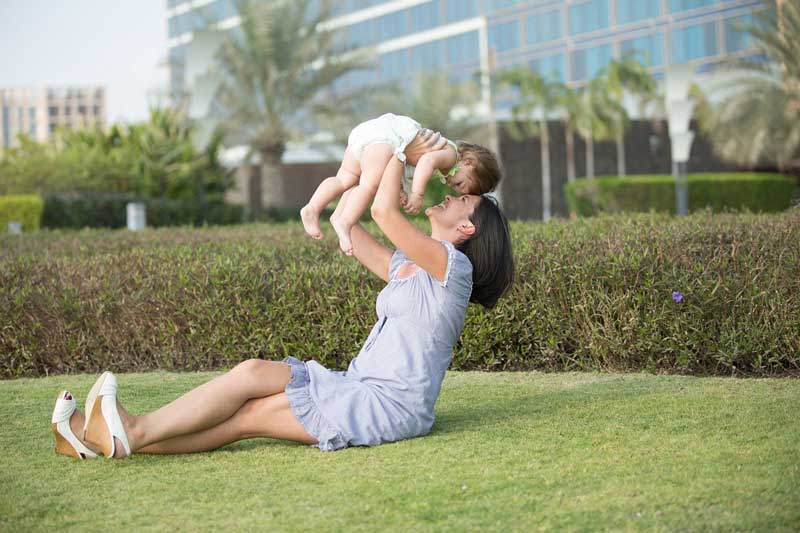 Frau auf Gras sitzend hebt Baby in die Luft mit Stadt im Hintergrund