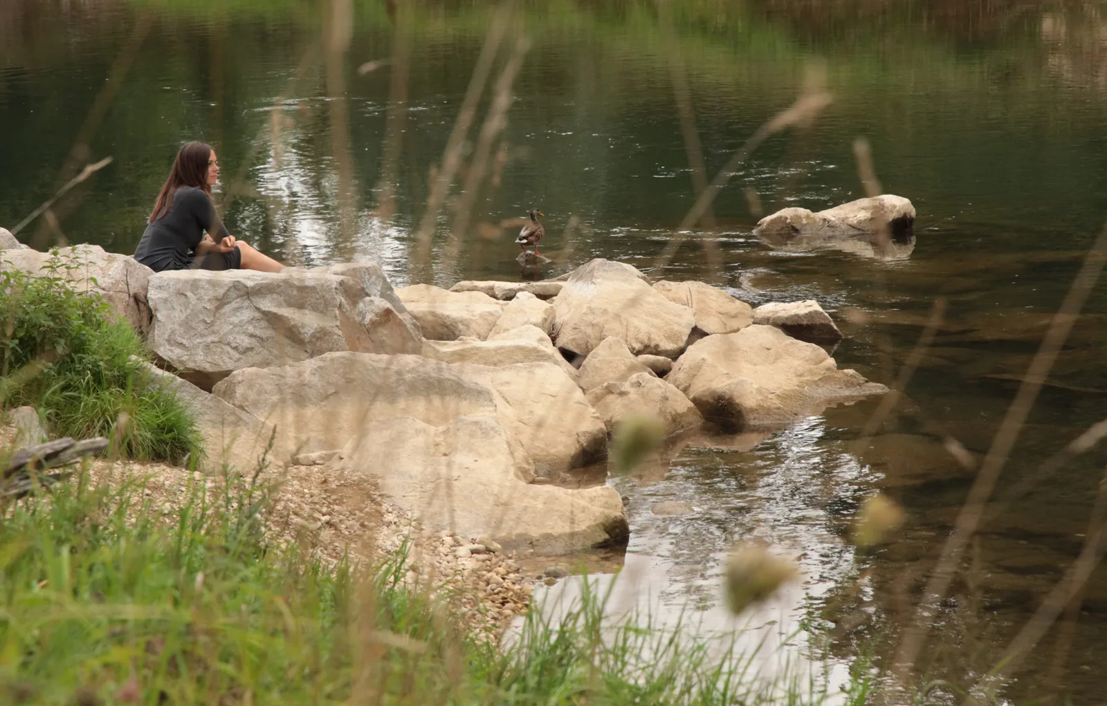 Frau mit rötlichen Haaren sitzt nachdenklich auf Steinen am Fluss mit Wildenten im Wasser