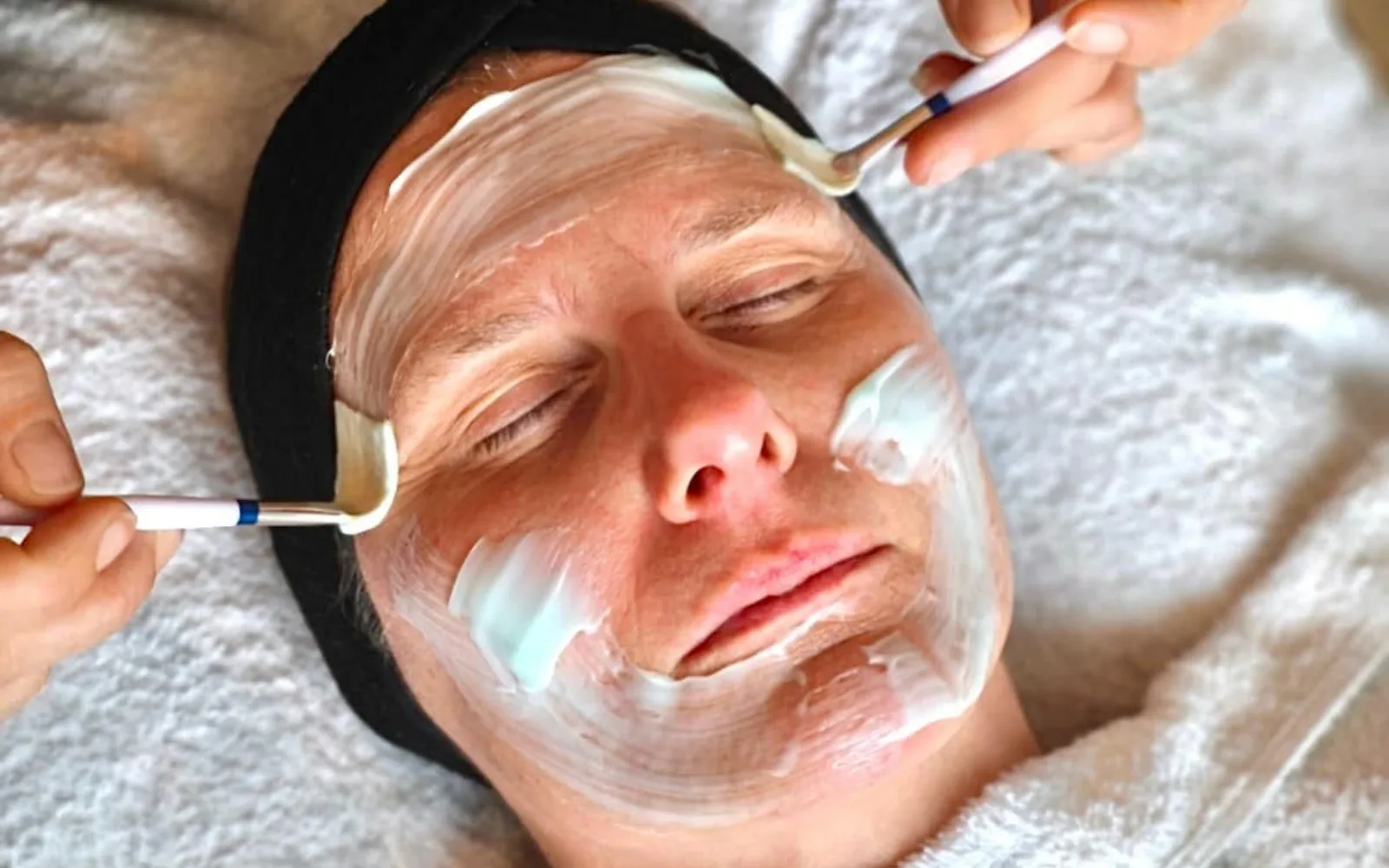 Frau mit Stirnband erhält Gesichtsbehandlung mit weißer Maske und Pinsel