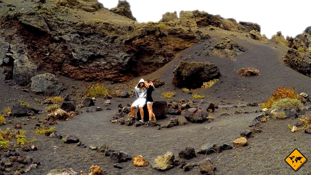Paar sitzt vor Vulkan El Cuervo im Timanfaya Nationalpark mit schwarzer Vulkanlandschaft