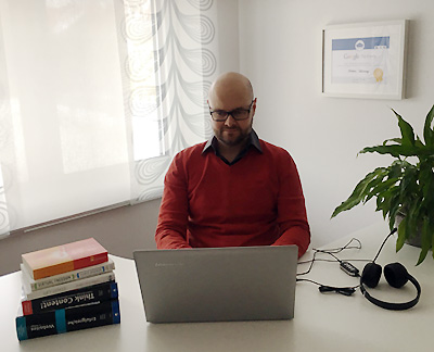 Mann mit Brille am Schreibtisch mit Laptop und Kopfhörern, Bücher und Pflanze im Hintergrund