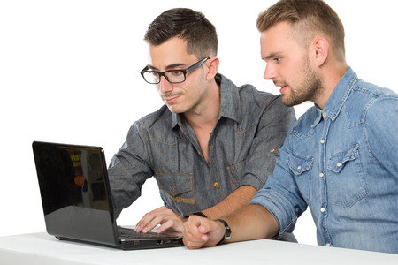 Zwei Männer schauen gemeinsam auf einen Laptop, beide tragen Hemden und sitzen an einem weißen Tisch