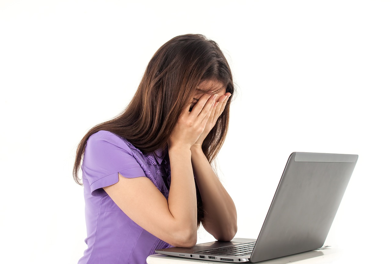 Junge Frau in lila Shirt sitzt frustriert am Laptop, Hand vor Gesicht, emotionaler Stress