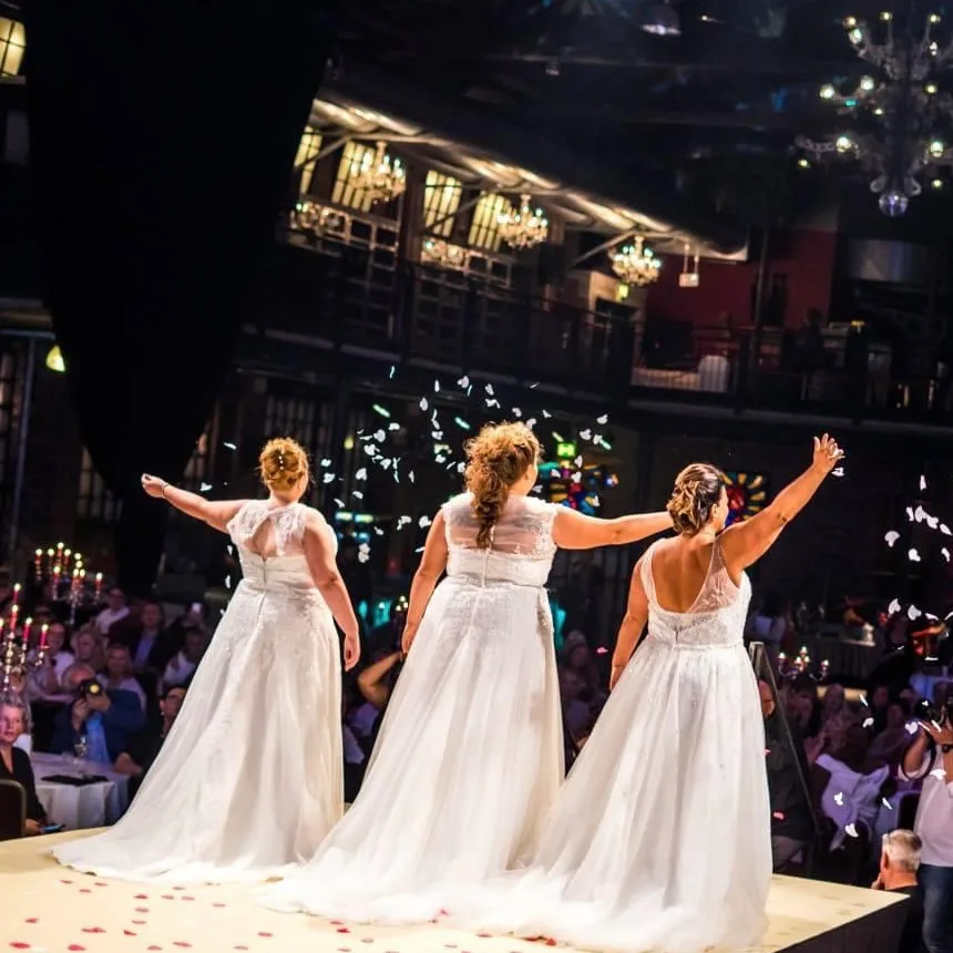 Drei Frauen in weißen Hochzeitskleidern auf Bühne mit ausgestreckten Armen, festliche Atmosphäre mit beleuchteter Kulisse