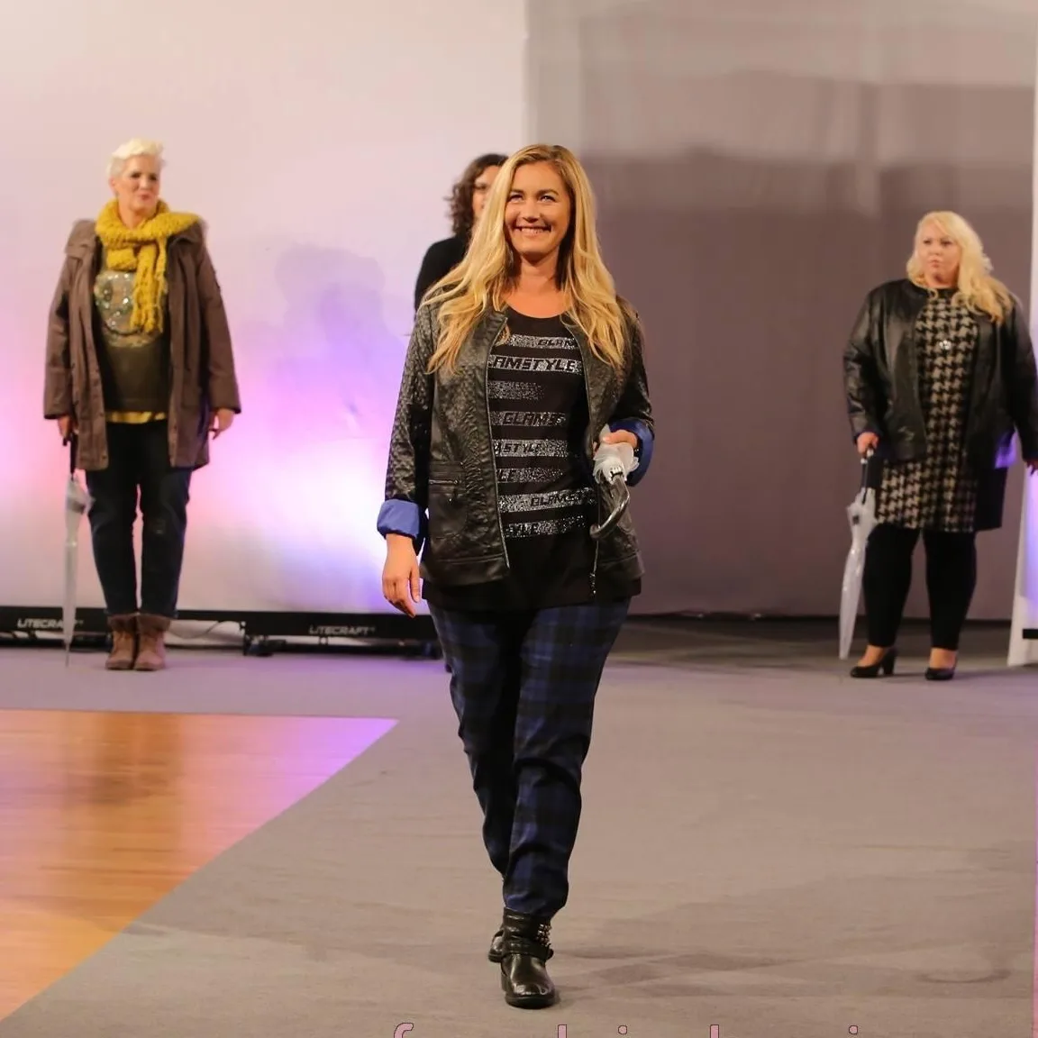 Frau in schwarzer Lederjacke und Jeans auf Laufsteg, zwei weitere Models im Hintergrund, rosafarbene Bühnenbeleuchtung