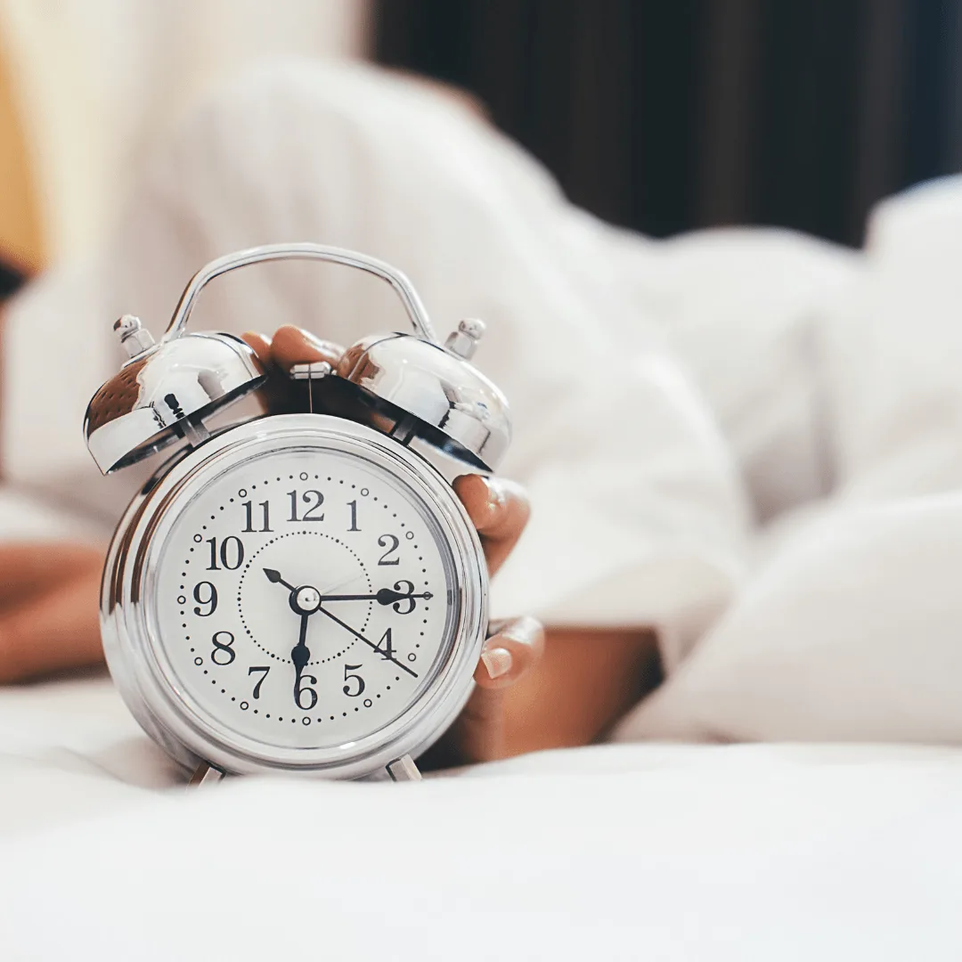 Adventskalender 2025 – Impulse bei Migräne und Stress 5 Silberne Wecker-Uhr auf weißem Bett mit Person im Hintergrund