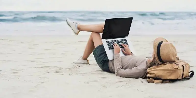 Person am Strand mit Laptop, Füße hochgelegt, entspanntes Arbeiten am Meer