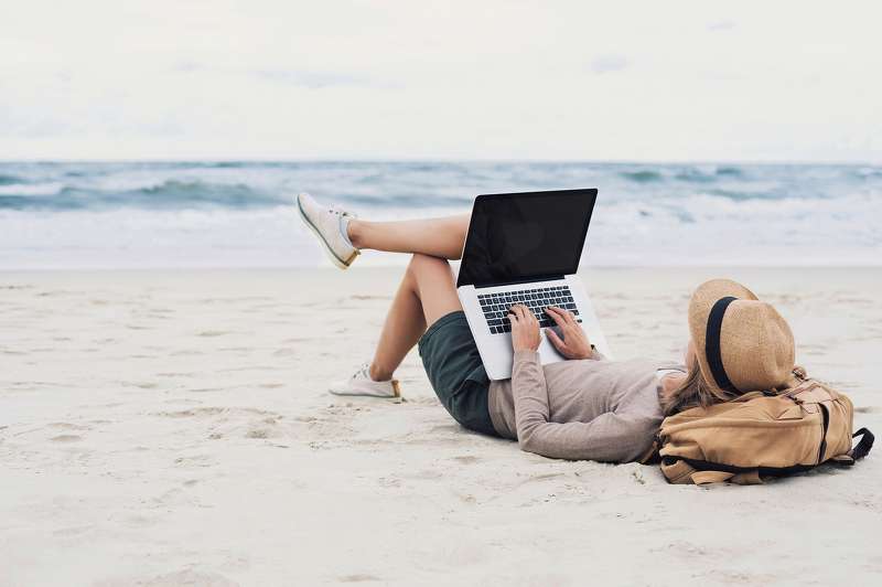 Person am Strand mit Laptop und erhobenen Beinen, arbeitet entspannt am Computer