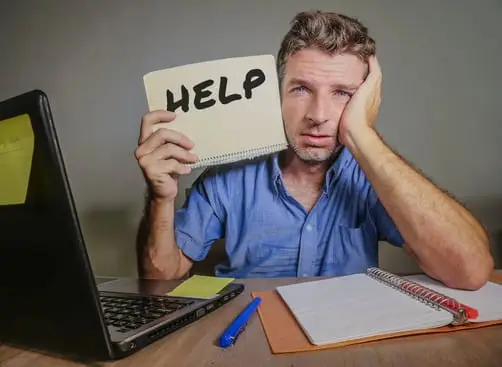 Mann am Schreibtisch vor Laptop hält Notizbuch mit Aufschrift HELP