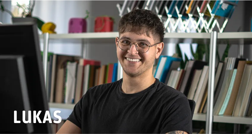 Portrait eines lächelnden Mannes mit Brille vor Bücherregal mit Text Lukas