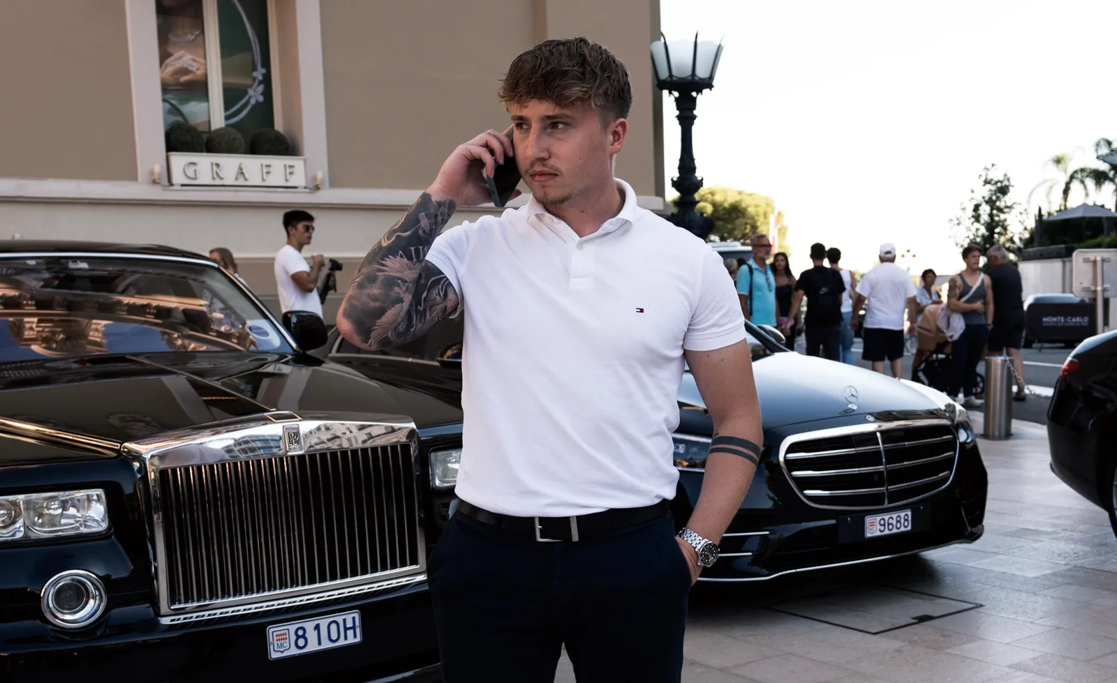 Mann in weißem T-Shirt telefoniert vor Luxusautos, Stadtplatz mit Publikum im Hintergrund