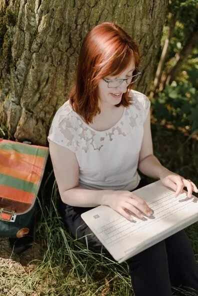 Frau mit rötlichem Haar und Brille sitzt lesend an Baum mit Laptop auf Schoß