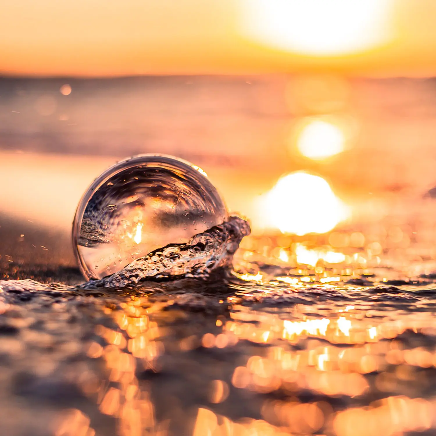 Glaskugel am Strand bei Sonnenuntergang, Wasser spritzt, goldene Lichter