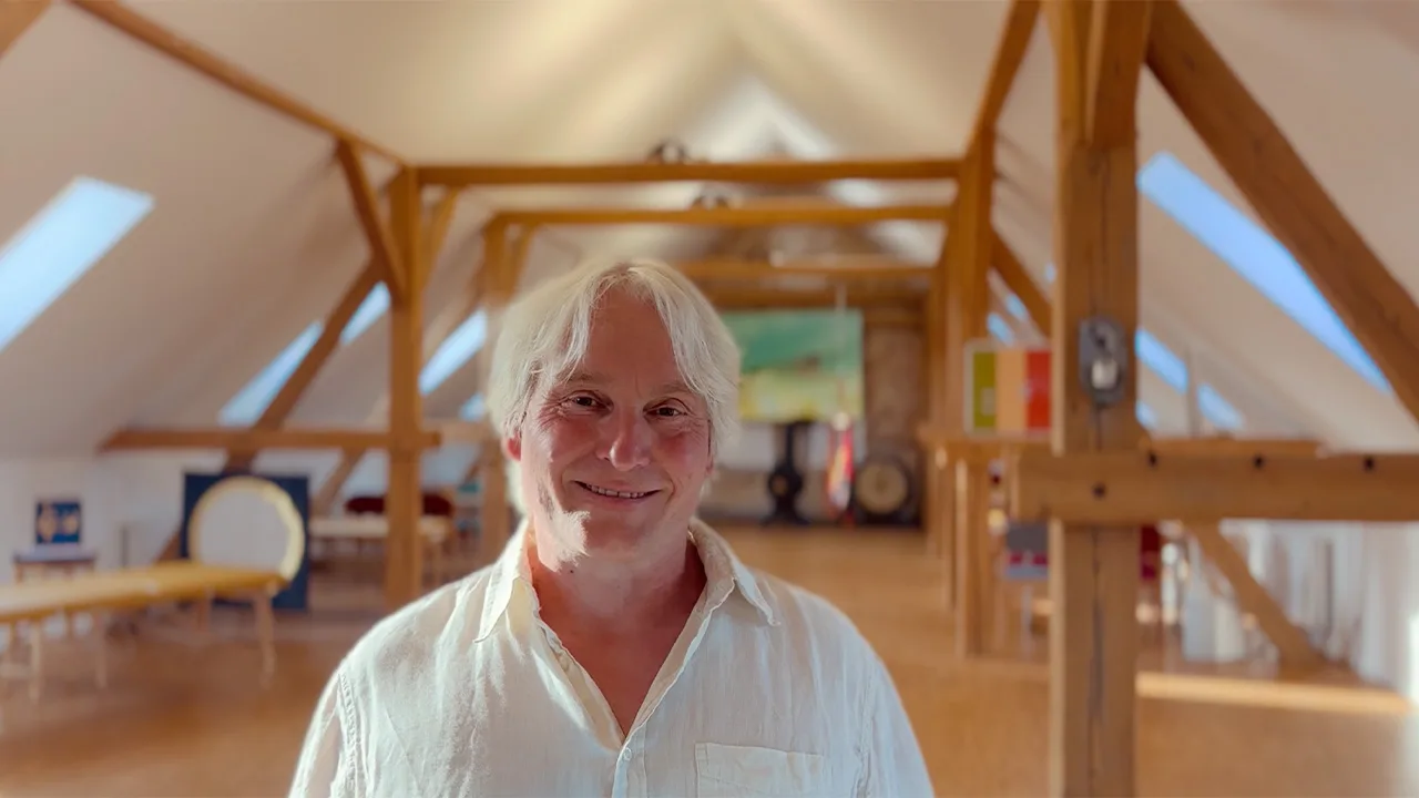 Mann mit weißen Haaren im hellen Hemd lächelt in einem modernen Holzgebäude mit Dachfenstern