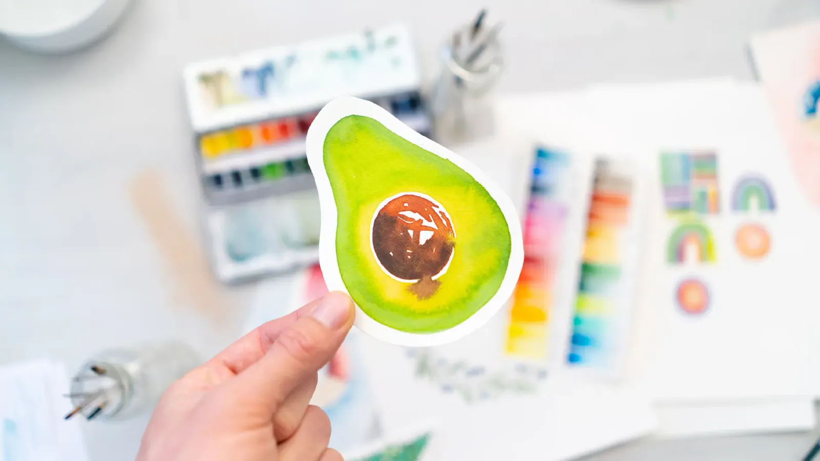 Aquarell-Skizze einer aufgeschnittenen Avocado mit grünem Fruchtfleisch und braunem Kern, gehalten in einer Hand vor unscharf