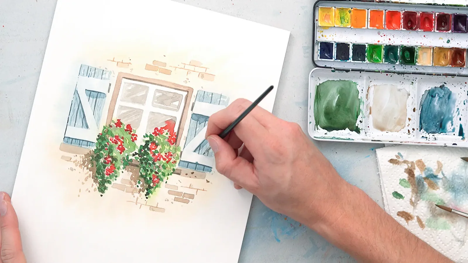 Hand mit Pinsel malt mehrschichtiges Aquarell eines Hauses mit roten Blüten an Fenster, professionelle Aquarellpalette danebe