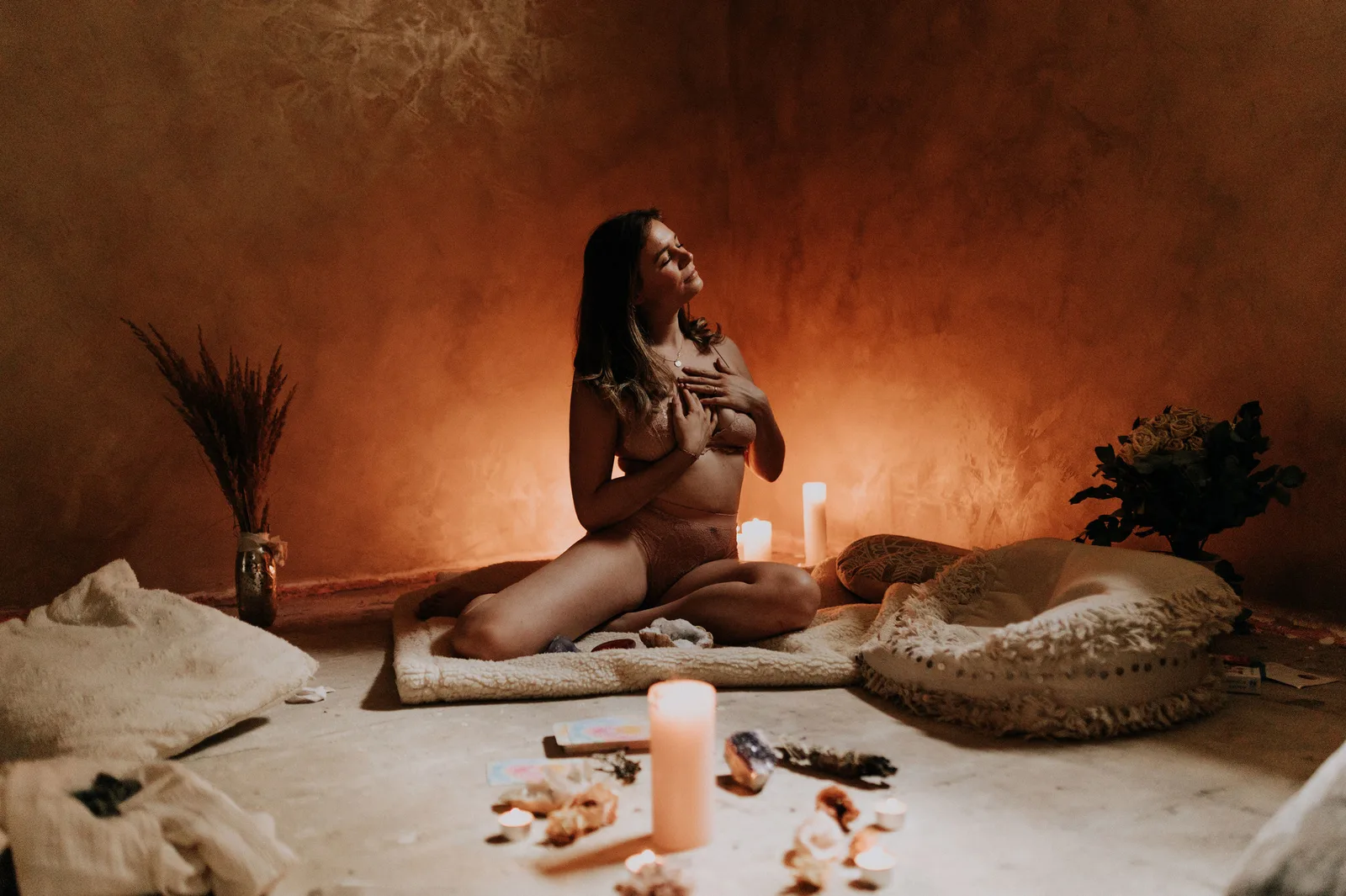 Frau in meditativer Pose auf Teppich mit Kerzen, Pflanzen und natürlichen Materialien in warmem orangebraunem Licht