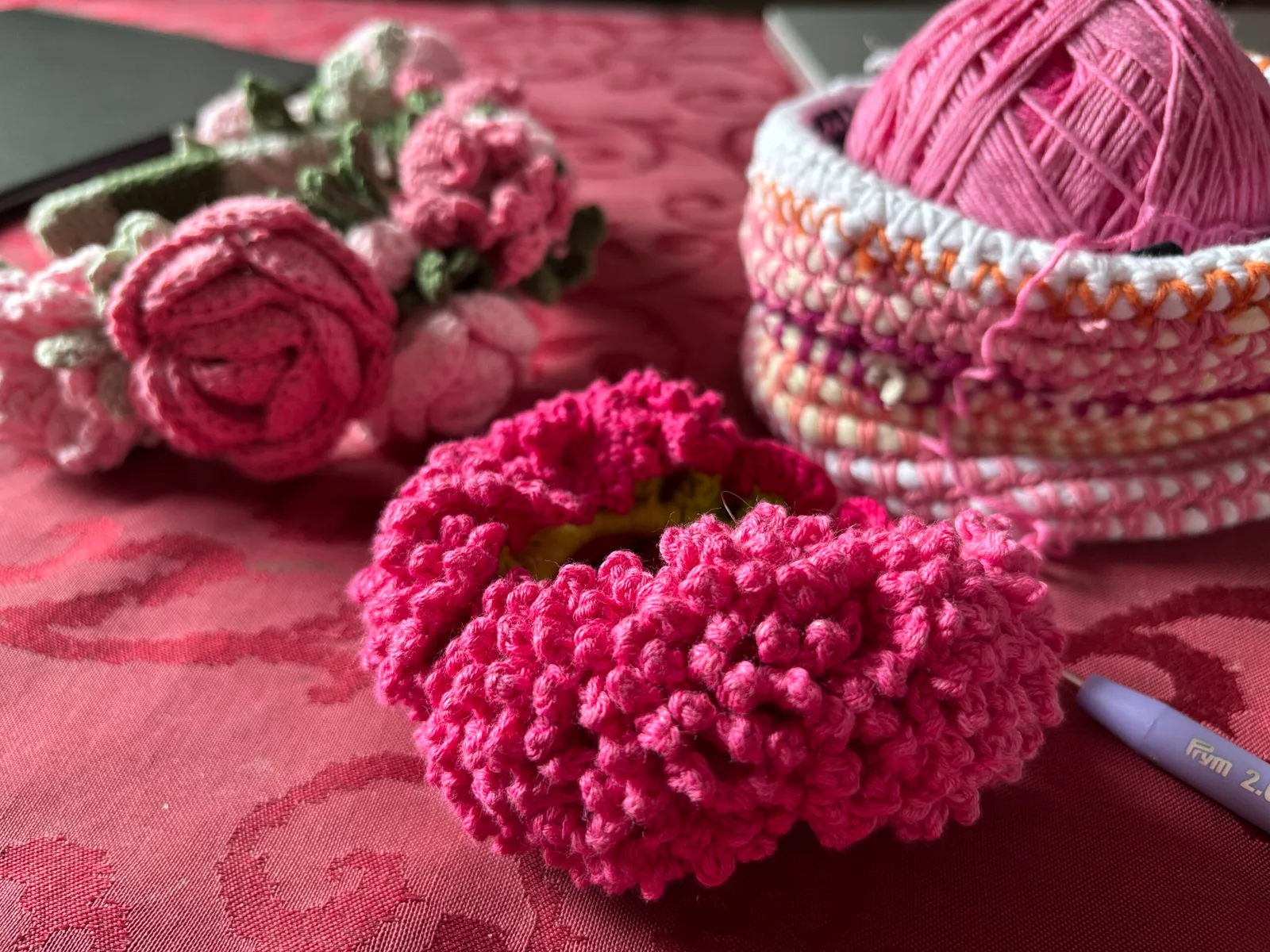 Gestrickte und gehäkelte Blüten in Pink und Magenta neben Wollknäueln und Häkelnadel auf rosa Stoff