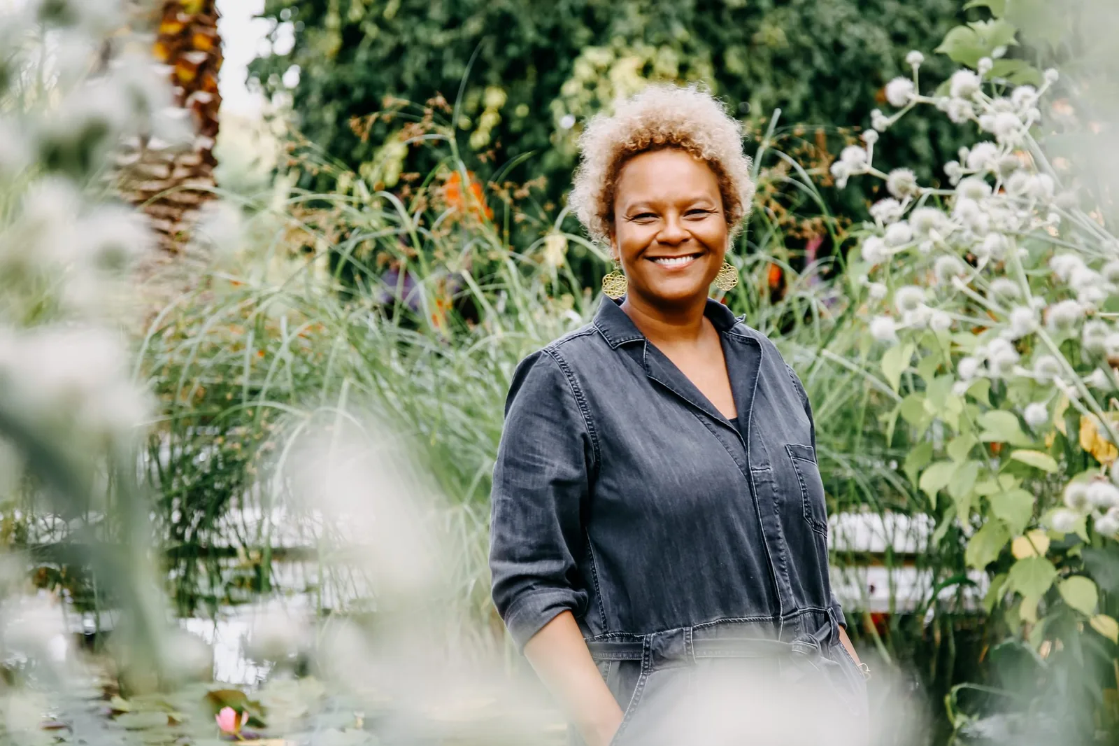 Frau mit weißem Haar und Denim-Hemd zwischen weißen Blüten und grünen Pflanzen, Garten