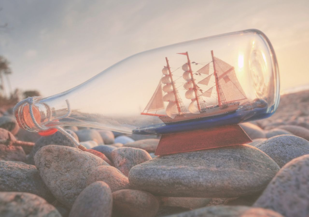 Segelschiff in Glasflasche auf Steinstrand bei Sonnenaufgang, symbolisiert Neubeginn und Abenteuer