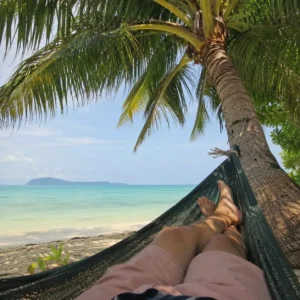 Person in Hängematte unter Kokospalme mit Blick auf türkisfarbenes Meer und Insel