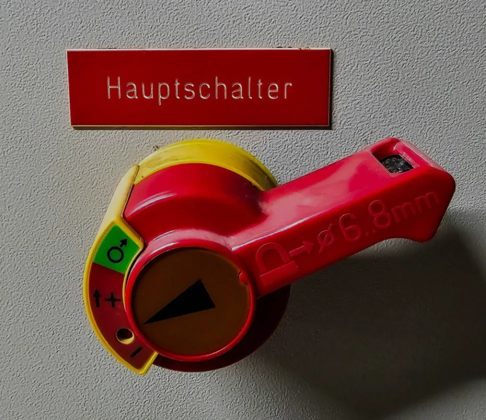Roter Hauptschalter mit Beschriftung an grauer Wand, gelb-grüne Markierung