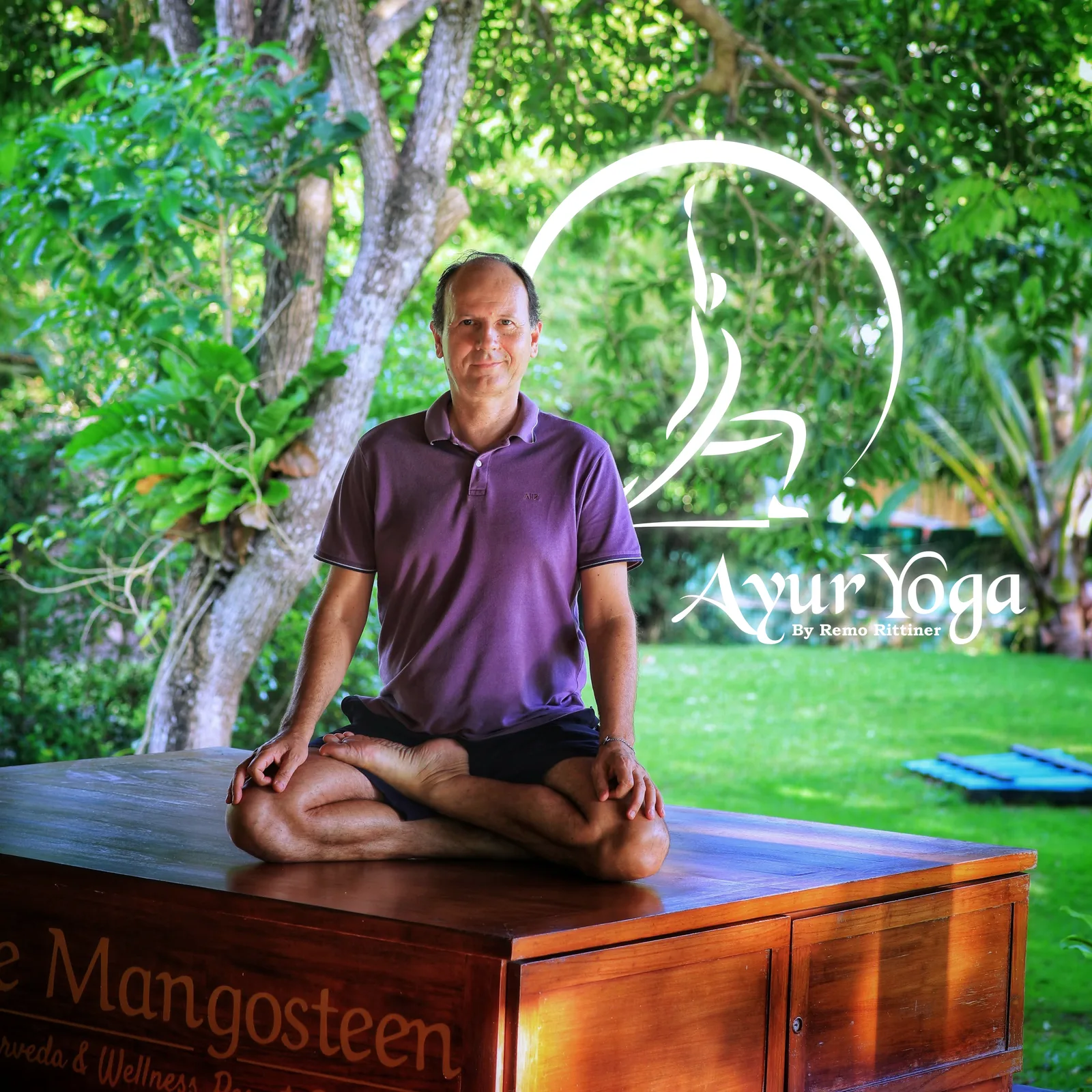 Remo Rittiner sitzt meditierend auf einem Holztisch im Garten, umgeben von grünem Laub und Bäumen, mit Ayur Yoga Logo