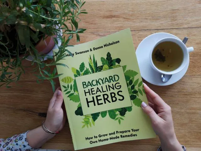 Backyard Healing Herbs Buch in Händen, daneben Tasse mit Tee auf Holztisch