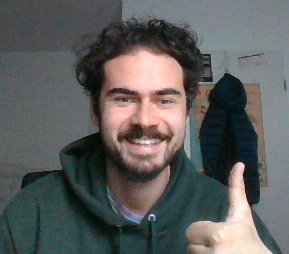 Junger Mann mit dunklen lockigen Haaren und Bart in grünem Hoodie, lächelnd, Daumen hoch