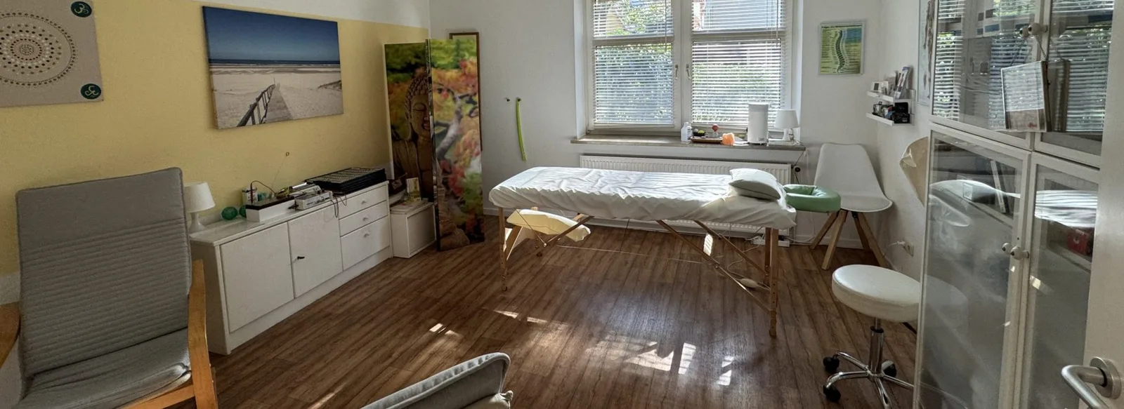 Praxis-Behandlungsraum mit Behandlungsliege, weißen Möbeln und großen Fenstern