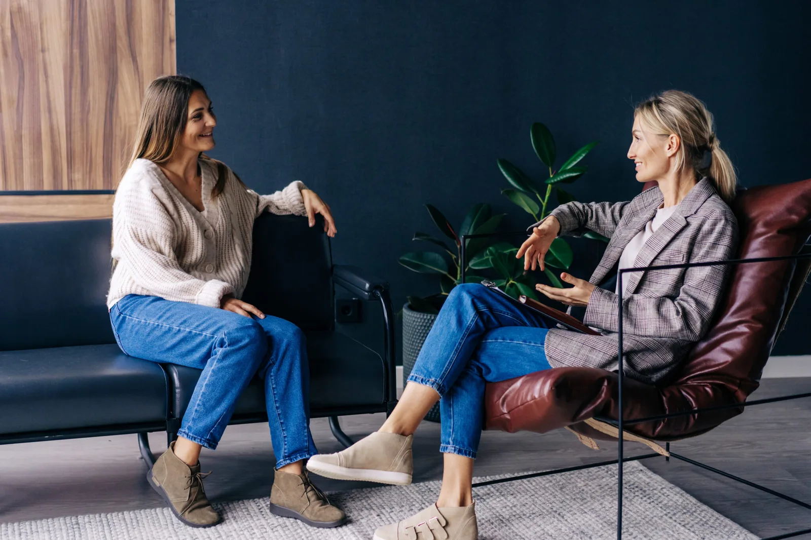 Zwei Frauen im Gespräch auf Sofa mit Pflanze und dunklem Hintergrund, lockere Coaching-Atmosphäre