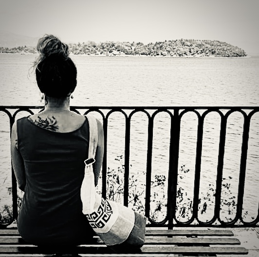 Frau sitzt an Geländer am Wasser und blickt auf Insel, Schwarz-Weiß-Fotografie