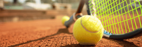 Tennisball auf Sandplatz neben Tennisschläger mit grünem Netz im Hintergrund