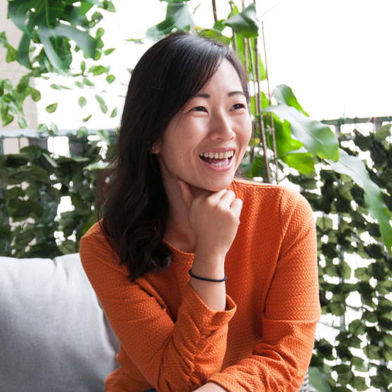 Portrait von Xiaoyu Dong, Frau in orangefarbenem Pullover vor Pflanzen