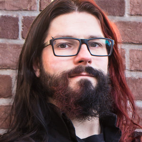 Portrait von Marek Firlej, Mann mit langen Haaren, Bart und Brille vor Backsteinwand