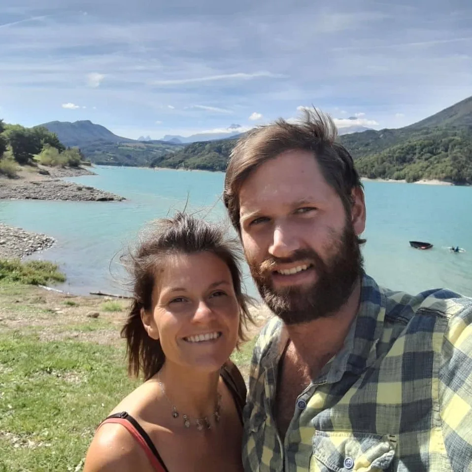 Lisa und Andy lächeln zusammen an türkisfarbenem Bergsee mit grünen Bergen im Hintergrund