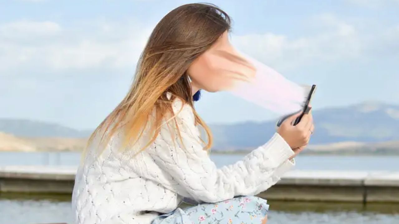 Junge Frau mit braunen Haaren hält Smartphone in die Luft, trägt weiße Jacke, Wasser und Berge im Hintergrund